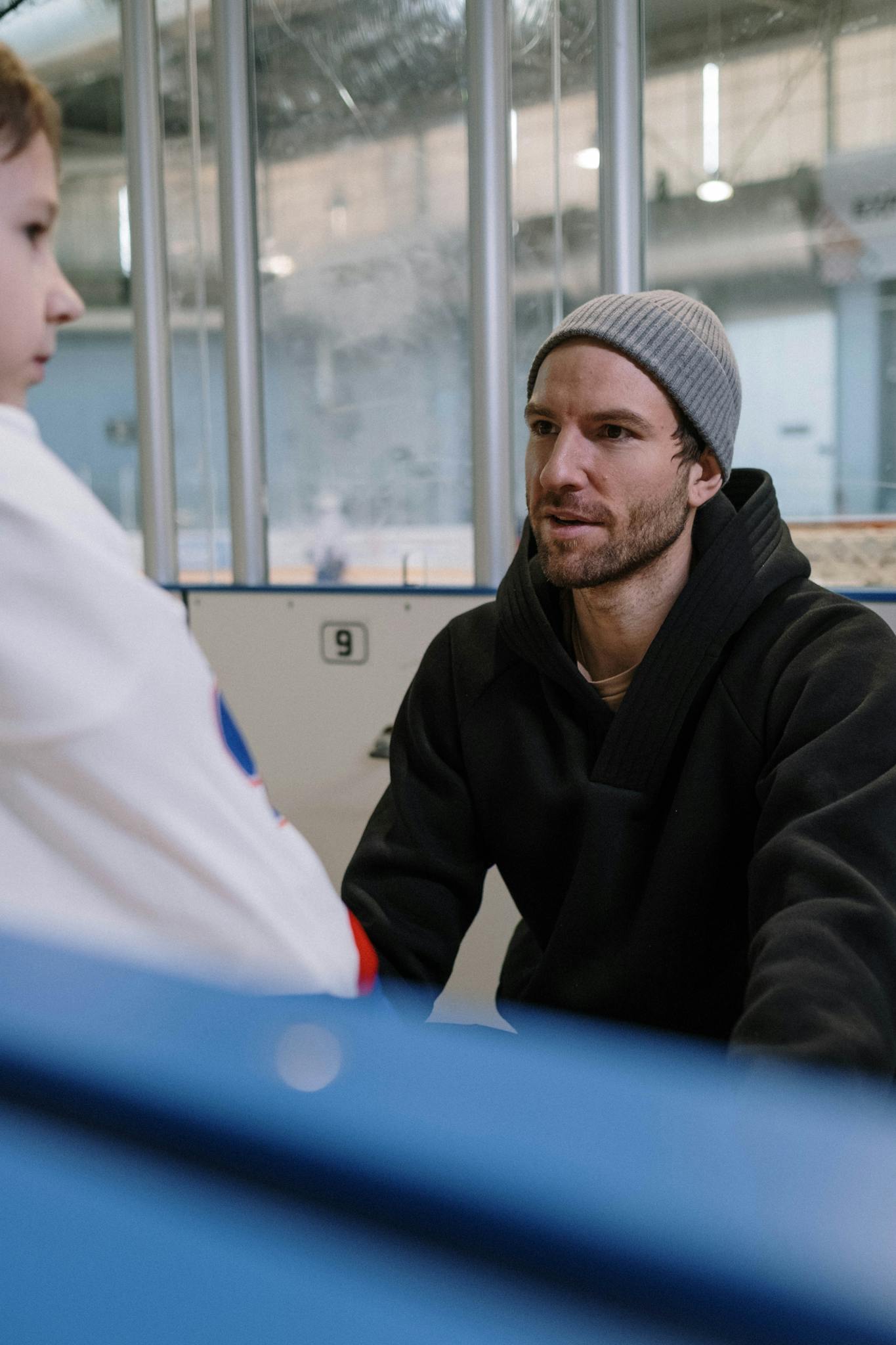 A father talks to his child at an indoor ice rink, wearing a beanie and winter clothes.